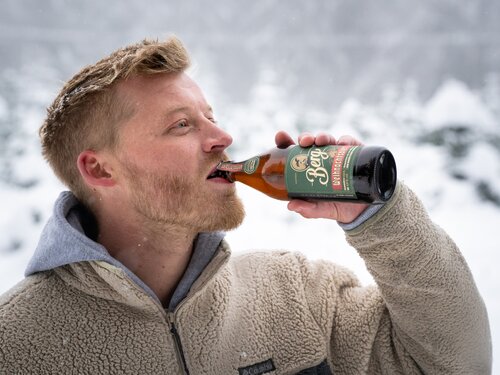 Ein Mann trinkt aus einer Bierflasche im Schnee, umgeben von einer winterlichen Landschaft.