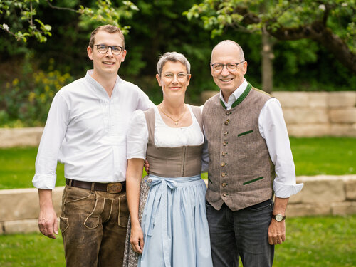 Eine Frau und zwei Männer in traditioneller bayerischer Tracht stehen zusammen und lächeln freundlich in die Kamera.